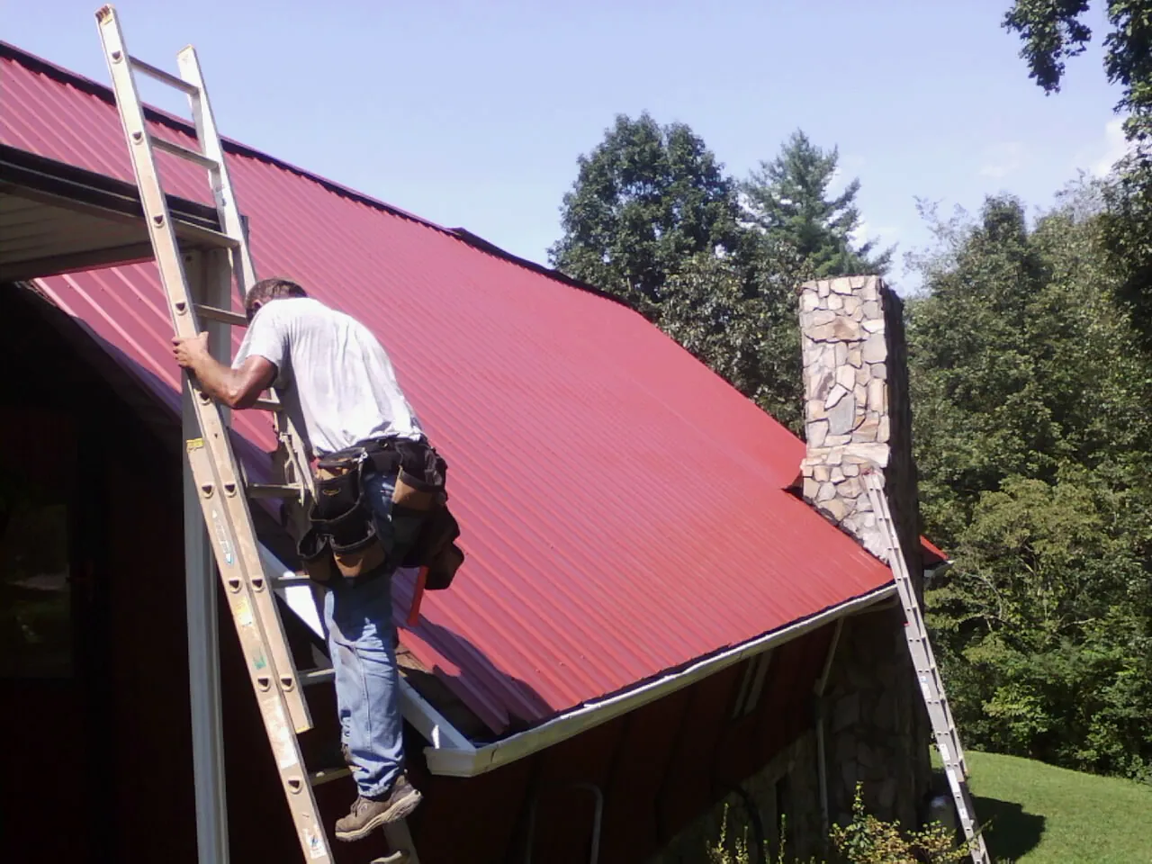 Roofer on a ladder beside a red metal roof and stone chimney — Soffit & Fascia Repair by Roofing Brothers in Peterborough